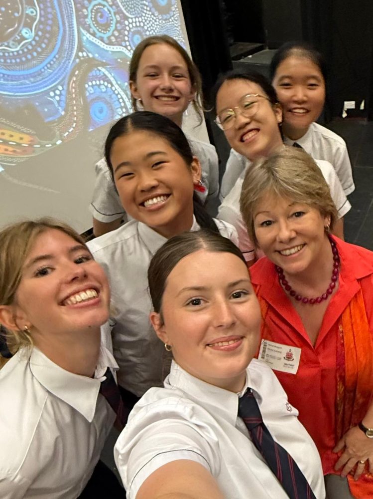 Students in school uniforms participating in an educational event promoting gender equality, posing with a presenter beside a projected cultural artwork